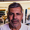 A close-up headshot of a mature man with graying hair and a mustache, looking directly at the camera with a steady, professional expression. He is wearing a white t-shirt, and the background features a blurred workshop wall organized with various hand tools, including scissors and chisels.