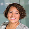 A close-up headshot of a smiling woman with short, curly brunette hair. She has a warm and welcoming expression and is wearing a gray-and-white striped blazer, set against a soft-focus office background with glass partitions.