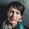 A close-up headshot of a woman with short, dark hair styled to the side. She is looking directly at the camera with a slight, confident smile. She is wearing large, circular silver earrings and a textured teal-and-white scarf, set against a dark, moody background with subtle lighting.