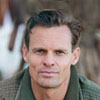 A close-up portrait of a mature man with brown hair and light eyes, looking directly at the camera with a calm and professional expression. He is outdoors, wearing a dark green textured sweater or jacket, with a soft-focus natural background.