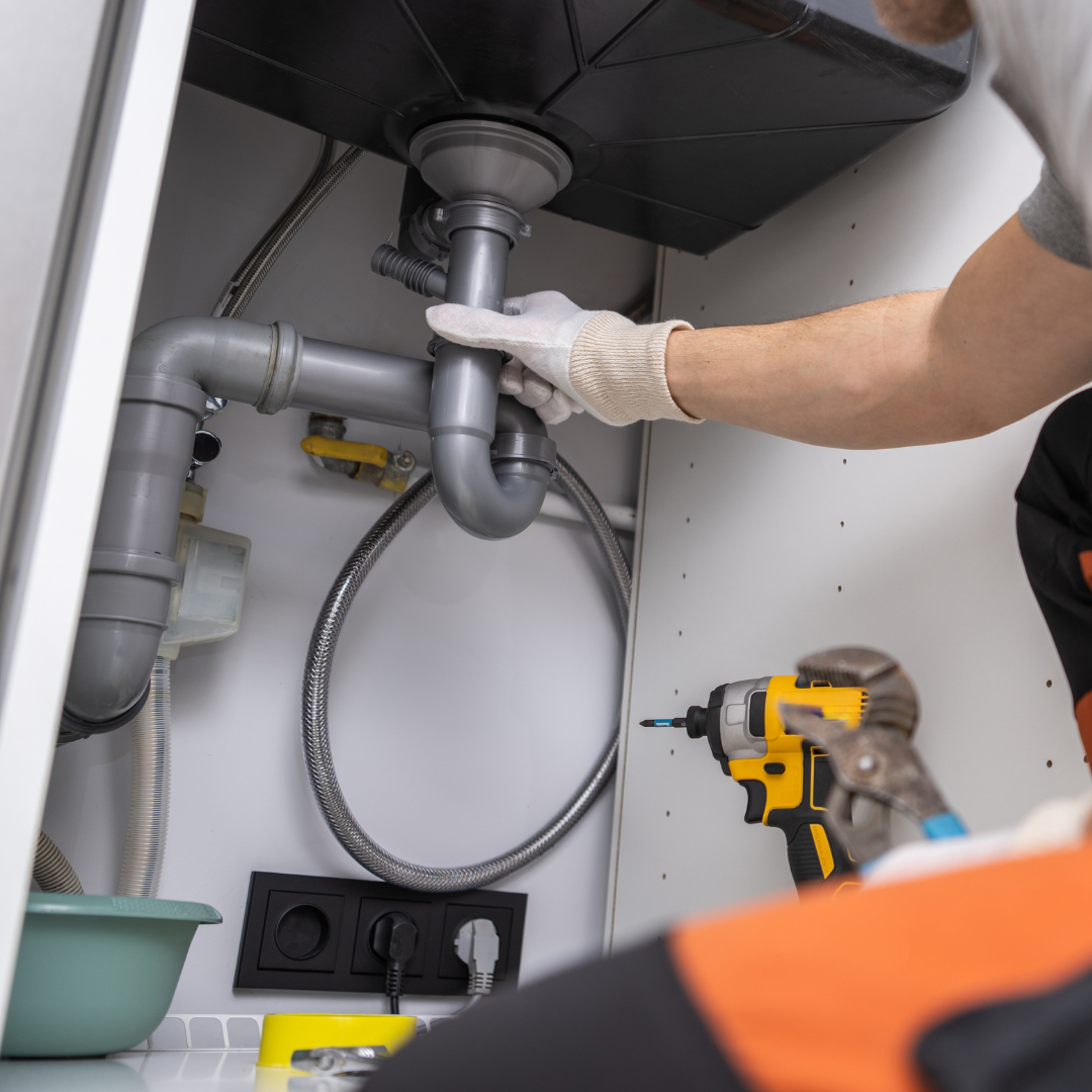 A plumber working under a kitchen sink, adjusting a grey plastic P-trap pipe. A yellow power drill and pliers are visible in the foreground on the cabinet floor.