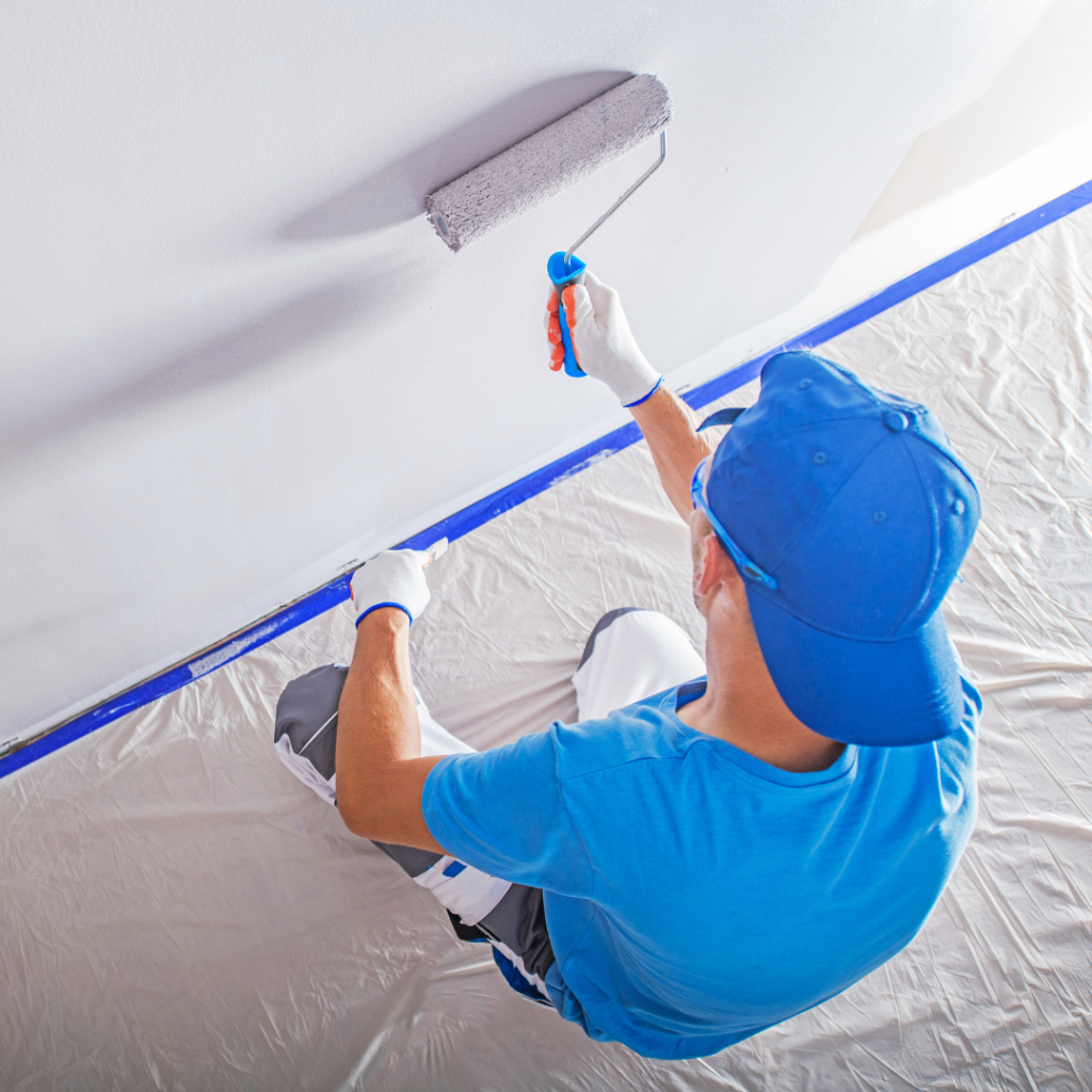 High-angle view of a painter in a blue shirt and cap using a roller to apply white paint to a wall. The baseboard is protected with blue painter's tape and the floor is covered with a plastic drop cloth.
