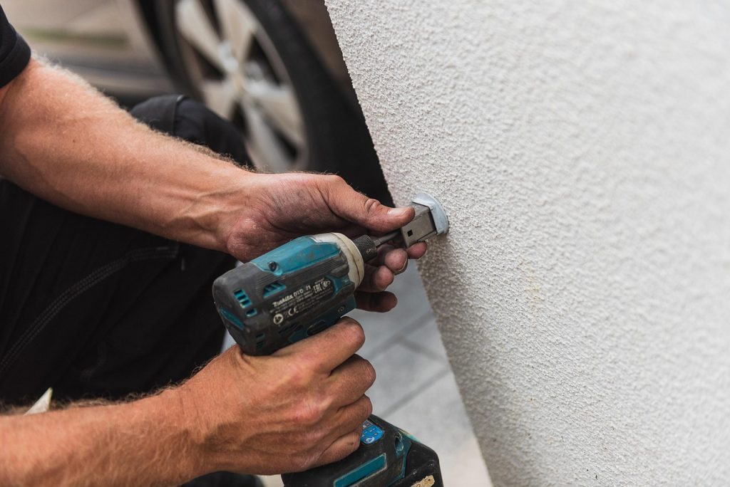 Close-up of a technician's hands using a blue Makita impact driver to secure a metal bracket or fixture into a white textured exterior wall.